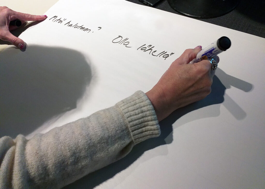 Käsi kirjoittaa suurelle paperille. Paperilla lukee "Mitä halutaan? Olla lähellä".