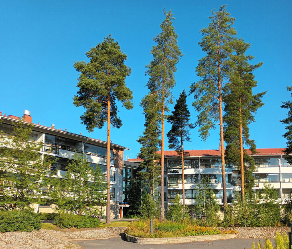 Kaksi kolmikerroksista lomahuoneistorakennusta. Mäntyjä ja nurmikkoa. Etualalla tie.