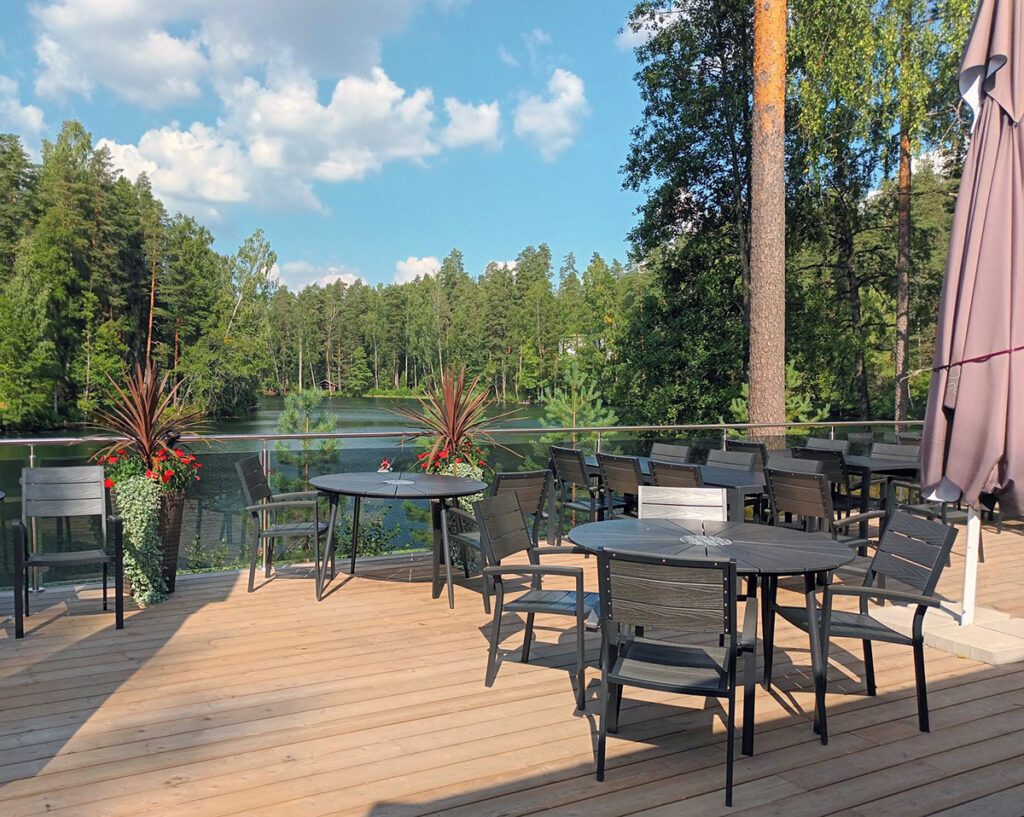 Ravintolan terassi, josta on näkymä järvelle. Vieressä kasvaa mäntyjä. Terassilla on kukka-asetelmia.