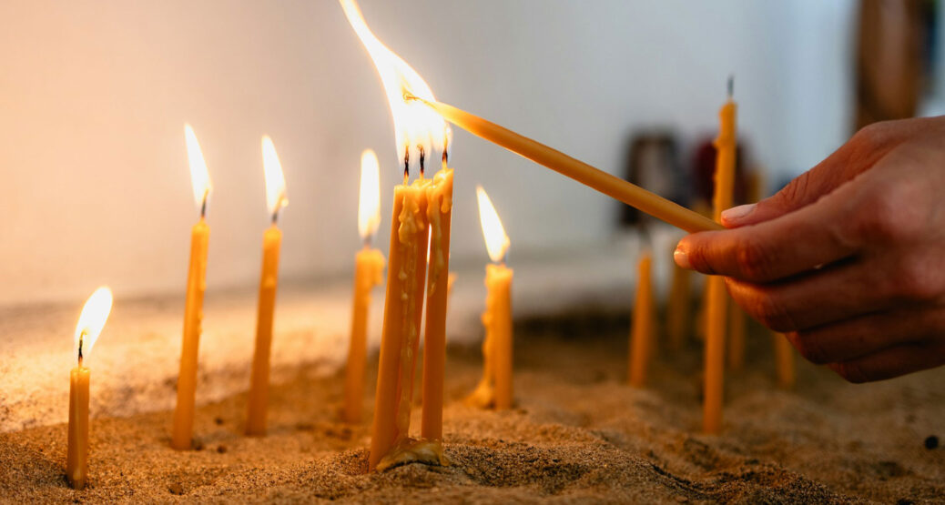 Hiekka-astiassa palavia tuohuksia. Etualalla tuohusta palavan tuohuksen liekistä sytyttävän henkilön käsi.