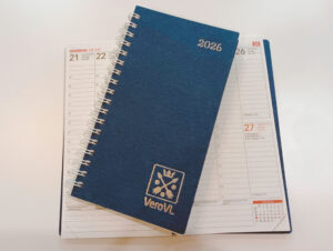 Kartonkikantinen taskukalenteri. Kalenterin kanteen on painettu hopeanvärisin kirjaimin 2026 ja VeroVL. Kalenterin alla on toinen kalenteri avonaisena. Auki olevasta kalenterista näkyy aukeama, jossa on yhden viikon päivät muistiinpanotiloineen.