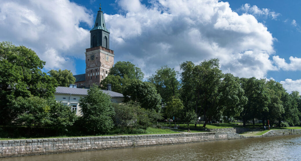 Aurajoki, jonka rannalla on Turun tuomiokirkko, puutalo ja vehreitä puita. On kesä. Kuva: Juho Luomala/Unsplash.